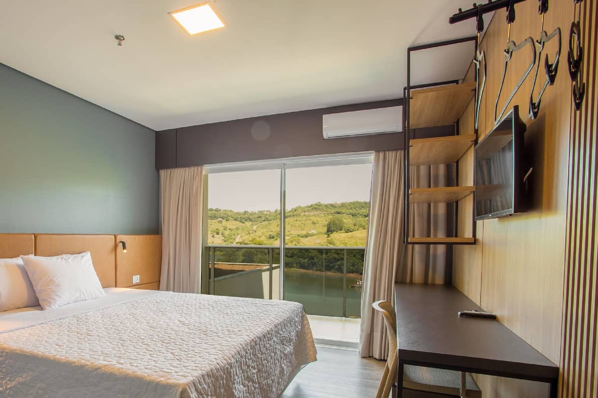A hotel room with a bed, a desk, a chair, a TV, and a balcony with a view of the landscape.