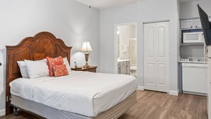 A bedroom with a wooden bed, white bedding, a nightstand with a lamp, and a view into a bathroom and kitchen area. - Queen Suite with Kitchenette (Branson)