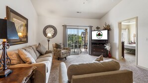 A living room with a sofa, armchair, and a television. There is a dining area with a table and chairs. The room has a ceiling fan and a large window.