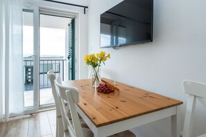 Un área de comedor con una mesa de madera, sillas blancas, un jarrón con flores amarillas y un televisor de pantalla plana montado en la pared.