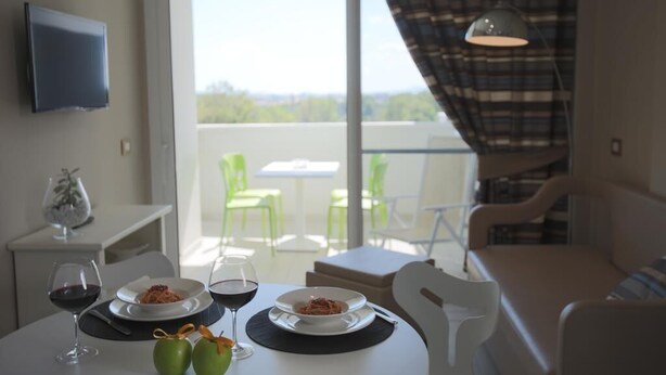 A dining area with a table set for two, two plates of food, two glasses of red wine, and two green apples. There is a television mounted on the wall and a window with a view of greenery outside. - 2-bedroom DELUXE apartment (Rimini)