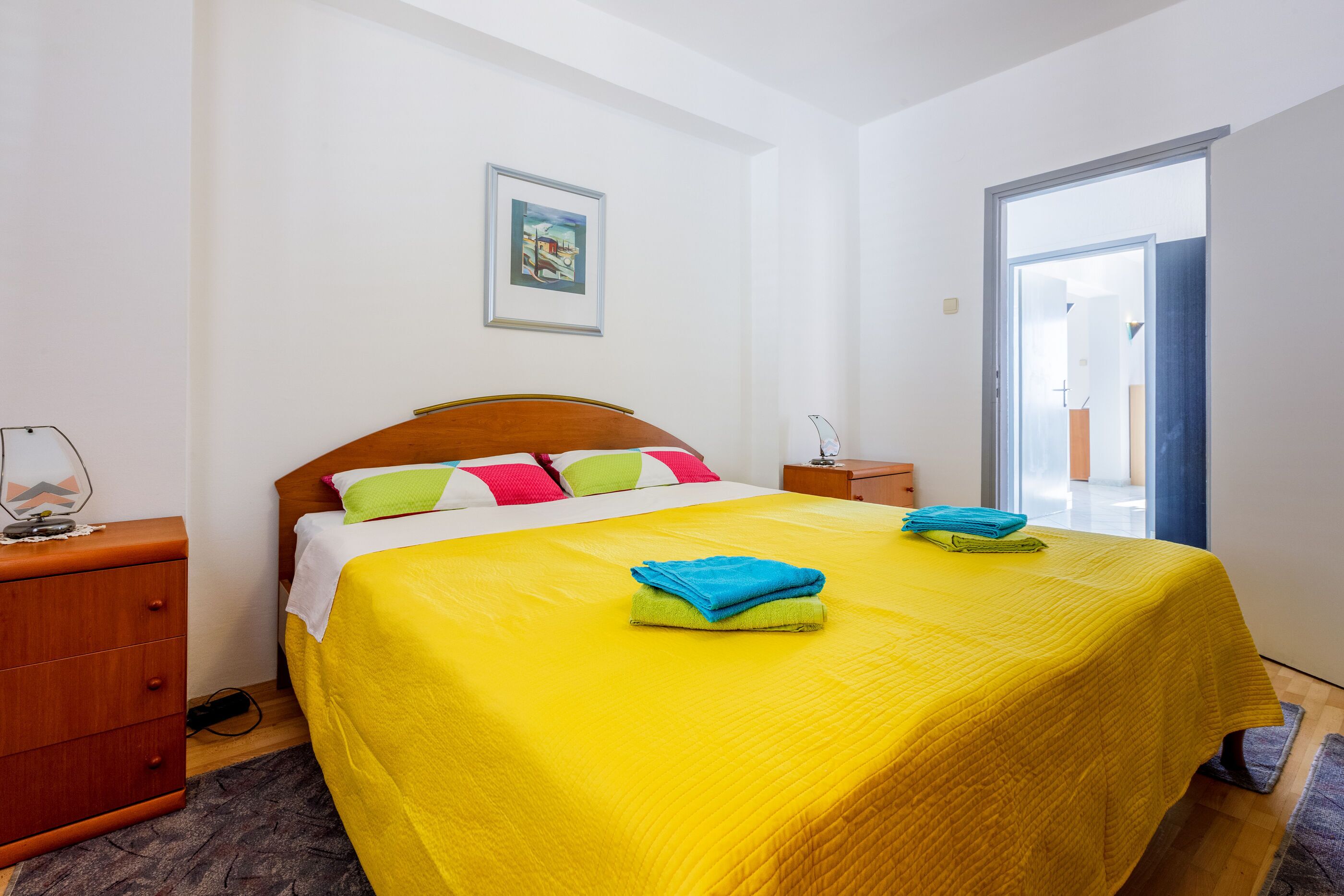 A neatly made bed with a yellow bedspread and colorful pillows, a wooden nightstand with a lamp, and a framed picture on the wall.