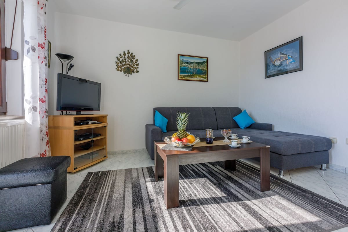 Una sala de estar con un sofá gris, una mesa de centro de madera con frutas, un televisor sobre un mueble de madera y un sillón negro.