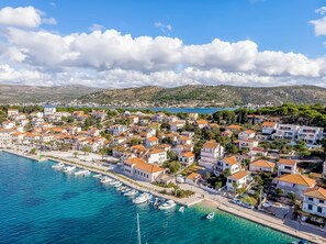 A coastal town with buildings along the waterfront, boats docked, and a clear view of the sea and hills in the background. - Apartments Cvitic (Rogoznica)