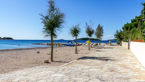 Un paseo marítimo empedrado con sillas y sombrillas de playa, que conduce a una playa arenosa con mar azul transparente y colinas al fondo.