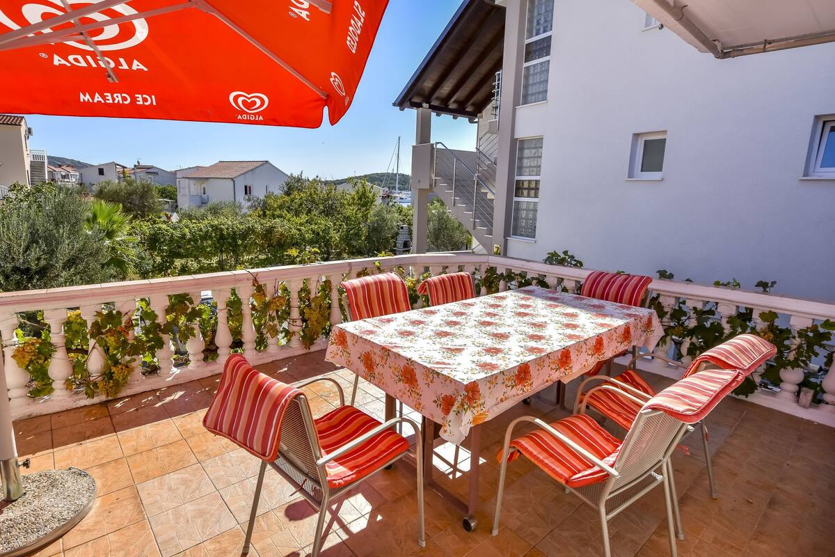 A balcony with a table and chairs, a red umbrella, and a view of buildings and greenery.