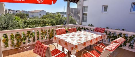 Un balcón con mesa y sillas, una sombrilla roja y vistas a edificios y vegetación.