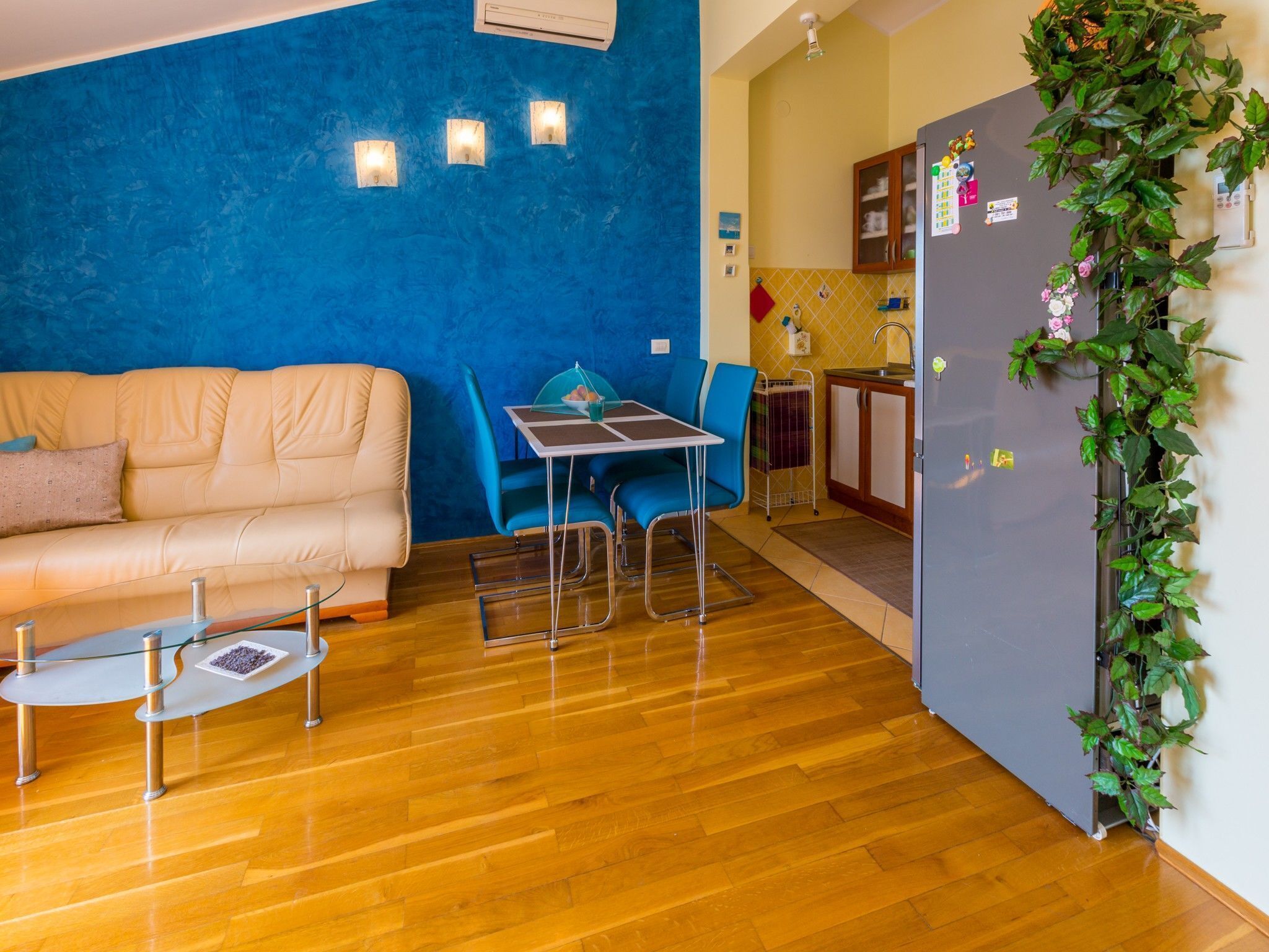Ein Wohnzimmer mit blauer Wand, einem beigen Sofa, einem Glastisch und einem Essbereich mit Tisch und Stühlen.