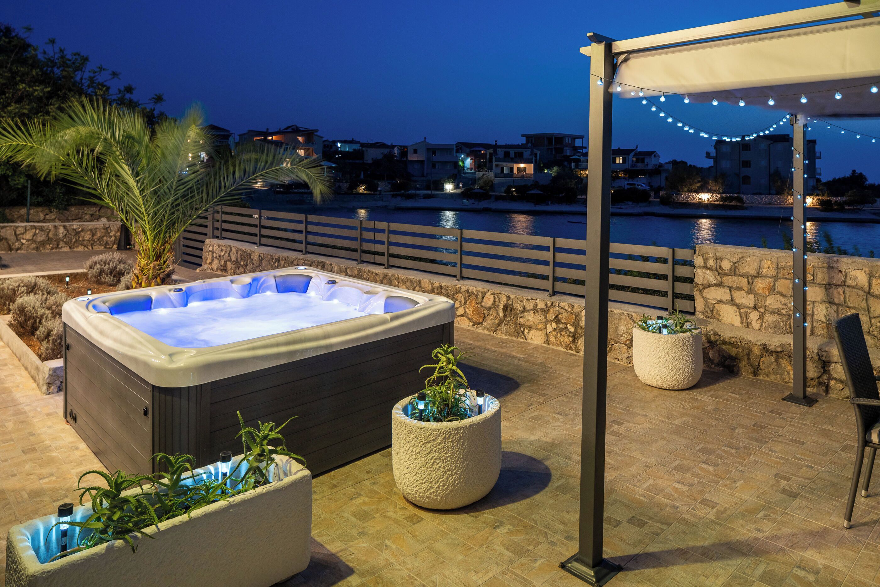 Ein Whirlpool, Kübelpflanzen, eine Pergola mit Lichterketten und Blick auf das Wasser.