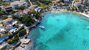 An aerial view of a coastal town with clear turquoise waters, boats, and buildings with red-tiled roofs.