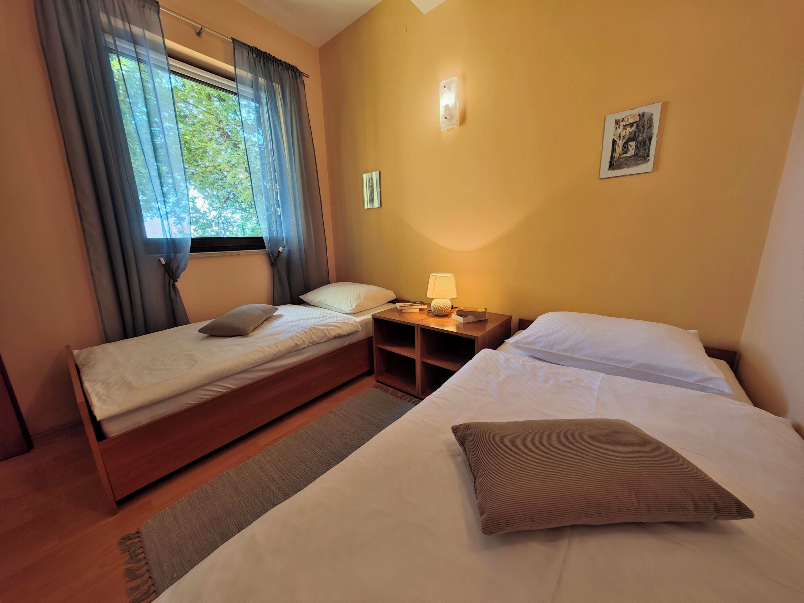 A hotel room with two beds, a nightstand with a lamp, a small shelf, and a window with curtains.