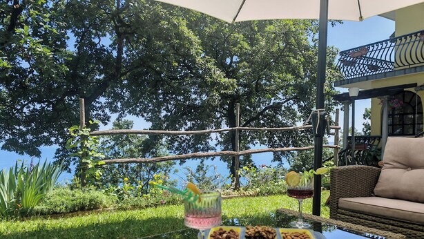 A glass-top table with food and drinks, wicker furniture, and a view of trees and a building. - Apartment Delost (Lovran)