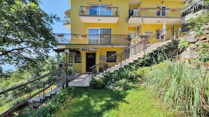 A multi-story yellow building with balconies, a staircase, and surrounding greenery. - Apartment Delost (Lovran)