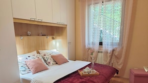 A neatly made bed with floral bedding, a red velvet bedspread, and a bedside table with a tray of tea and cookies. - Apartment Delost (Lovran)