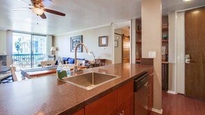 A modern kitchen with a granite countertop, stainless steel sink, and wooden cabinets. There is a ceiling fan and recessed lighting. - Beachfront 1BR Condo | Pool | Hot Tub | Sleep 4 (Oceanside)