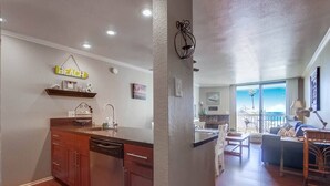 A kitchen with wooden cabinets, a dishwasher, and a countertop. There is a 'BEACH' sign on the wall and a window overlooking a balcony. - Beachfront 1BR Condo | Pool | Hot Tub | Sleep 4 (Oceanside)