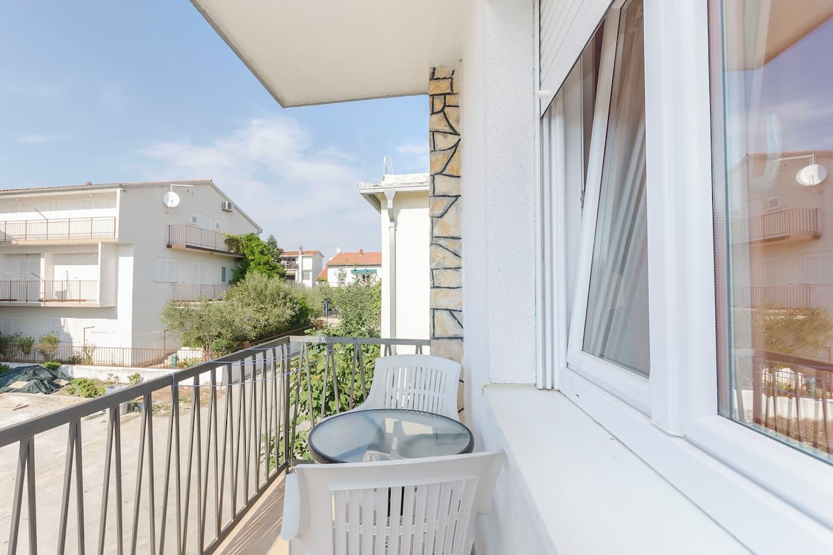 A balcony with a glass table and white chairs, overlooking a residential area with houses and a clear sky.