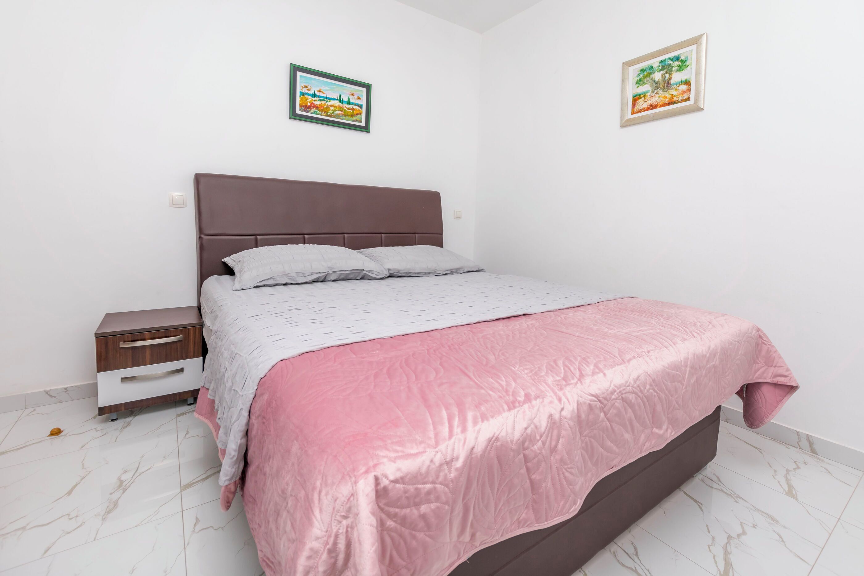 A bedroom with a bed, a nightstand, a drawer unit, and two framed pictures on the wall.