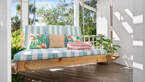 Eine Hängematte aus Holz mit gestreiften Kissen und blumigen Zierkissen, auf einer Holzterrasse unter einem weißen Pergola.