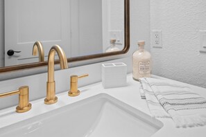 Une salle de bain avec un lavabo blanc, des robinets dorés, un grand miroir et un flacon de sels de bain.