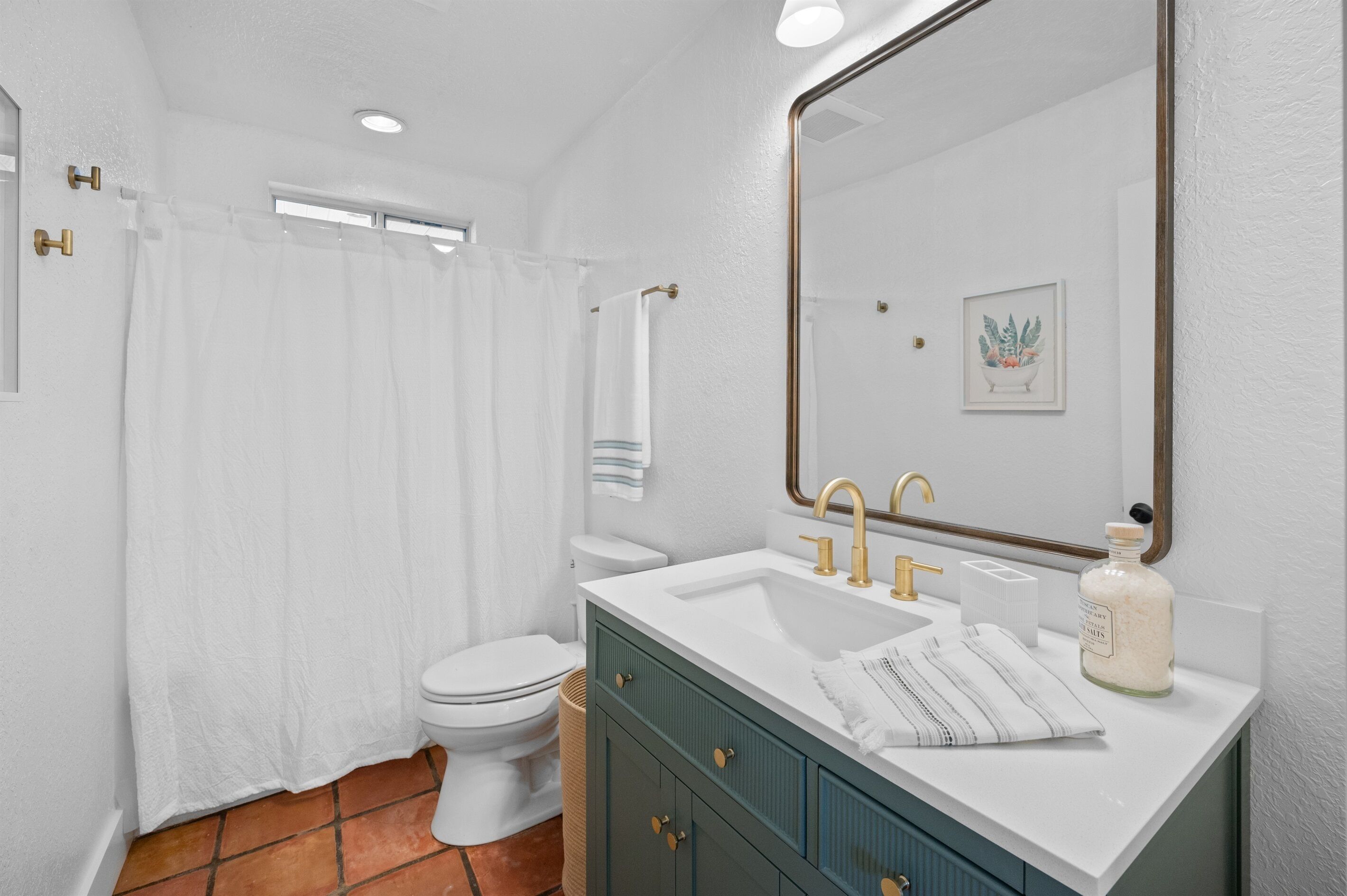Une salle de bain avec des toilettes blanches, une douche avec un rideau blanc, un lavabo avec un miroir et un meuble avec une serviette.