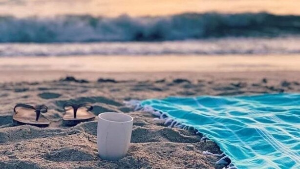 Eine Strandansicht mit einem Paar Flip-Flops und einer weißen Tasse im Sand, einem blauen Handtuch und dem Meer im Hintergrund.