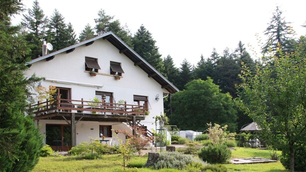 Une maison à deux étages dotée d’un balcon, entourée d’un jardin bien entretenu où poussent différentes espèces de plantes et où se trouve un petit étang.