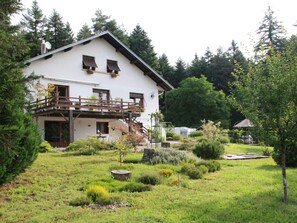Ein zweigeschossiges Haus mit Balkon, umgeben von einem gepflegten Garten mit verschiedenen Pflanzen und einem kleinen Teich.