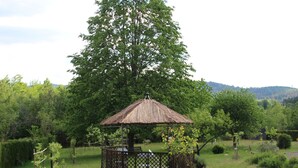 Un belvédère rustique, doté d’un toit de chaume, entouré d’une végétation luxuriante et d’arbres, dans un cadre extérieur paisible.