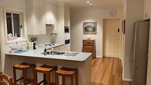 A modern kitchen with white cabinets, a central island with stools, a refrigerator, and a clock on the wall. - ☘️"KILMEEDY" Take time to smell the roses🌹 in Bowral-Walk to town-Pet friendly. (Bowral)