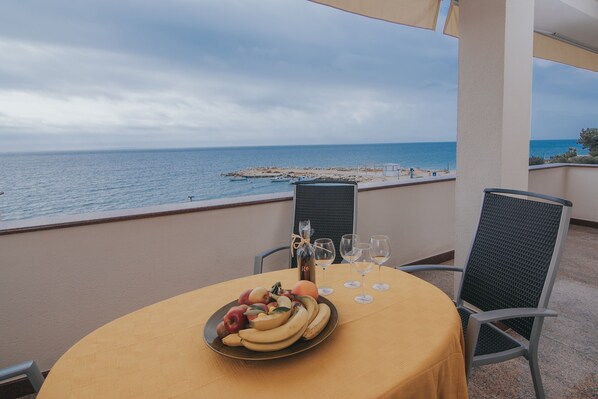 A table with a fruit platter, wine glasses, and a bottle on a balcony overlooking the sea.