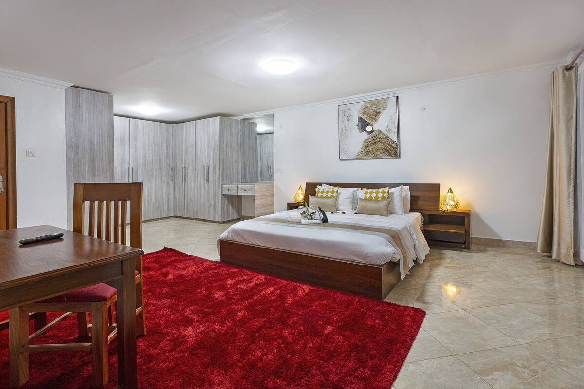 A bedroom with a bed, a wooden nightstand, a red rug, a wooden chair, and a wooden table.