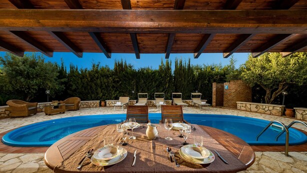 An outdoor pool area with a wooden table set for dining, surrounded by lounge chairs and a stone wall.