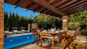 An outdoor pool area with a wooden pergola, lounge chairs, and a dining table set for six.