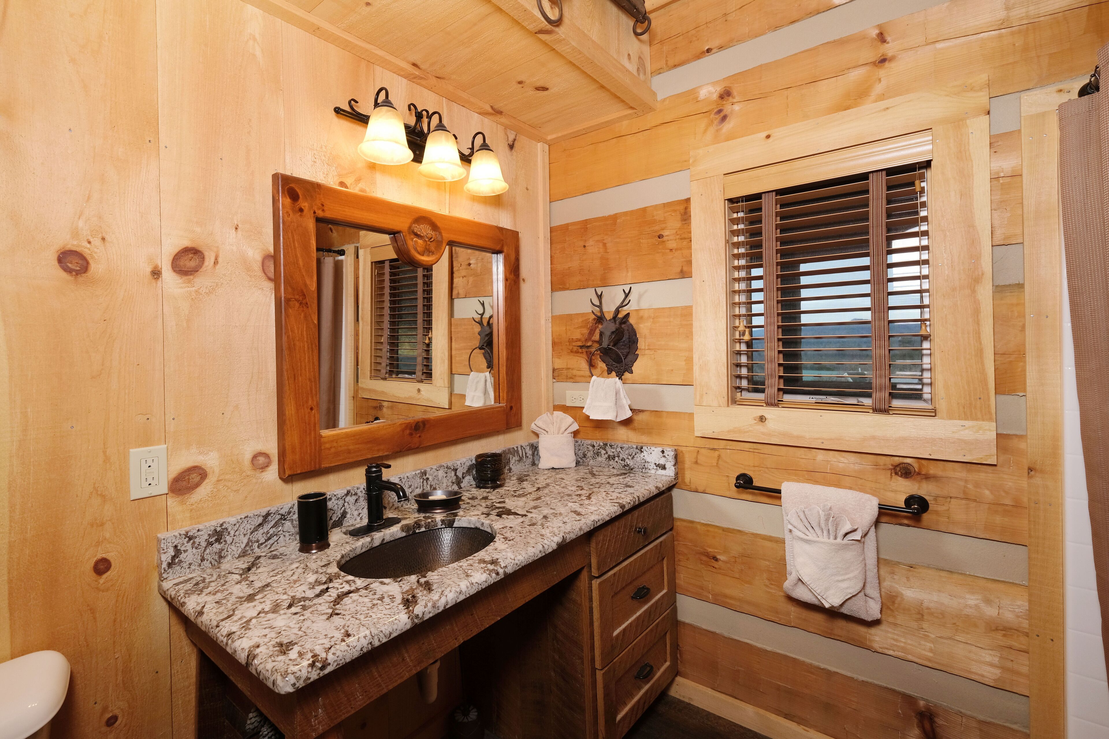 Une salle de bain rustique, avec une cabane en bois, un lavabo avec un plan de travail en granit, un miroir avec éclairage intégré, une fenêtre avec stores et un porte-serviettes.