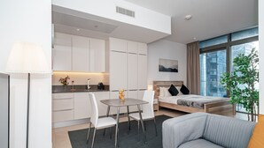 A modern hotel room with a bed, a gray sofa, a dining table with chairs, a kitchen area, and a large window with a view. - HiGuests - Marina Gate Tower (Dubai)