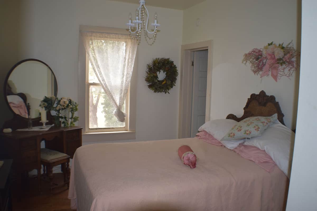 A bedroom with a bed, a dresser, a mirror, a window with curtains, a chandelier, and a wall decoration.