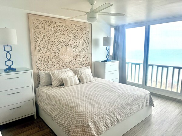 Un dormitorio con una cama grande, un cabecero decorativo, una mesita de noche, una lámpara y vista al mar a través de una ventana.