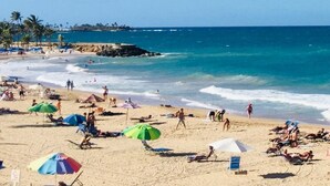 A beach with people sunbathing and swimming, umbrellas, and beach chairs. - The Happy Place (San Juan)