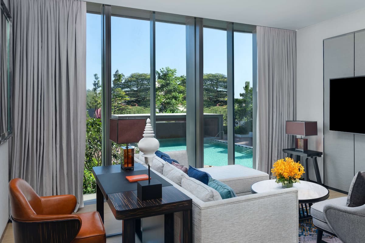 A modern hotel room with a large window, a flat-screen TV, a sofa, a coffee table, and a small table with a lamp and decorative items.