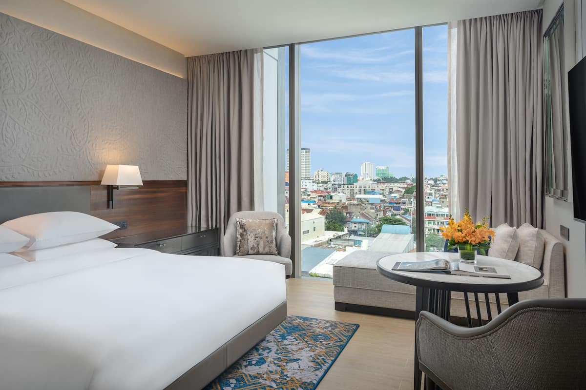 A hotel room with a large bed, a sofa, a small table, and a view of the city through large windows.