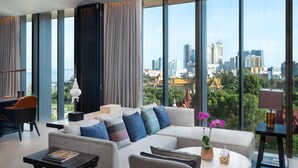 A modern hotel room with a large window offering a city view, a sofa with multiple cushions, a small table with a book and a vase of flowers, and a desk with a lamp. - Hyatt Regency Phnom Penh (Phnom Penh)