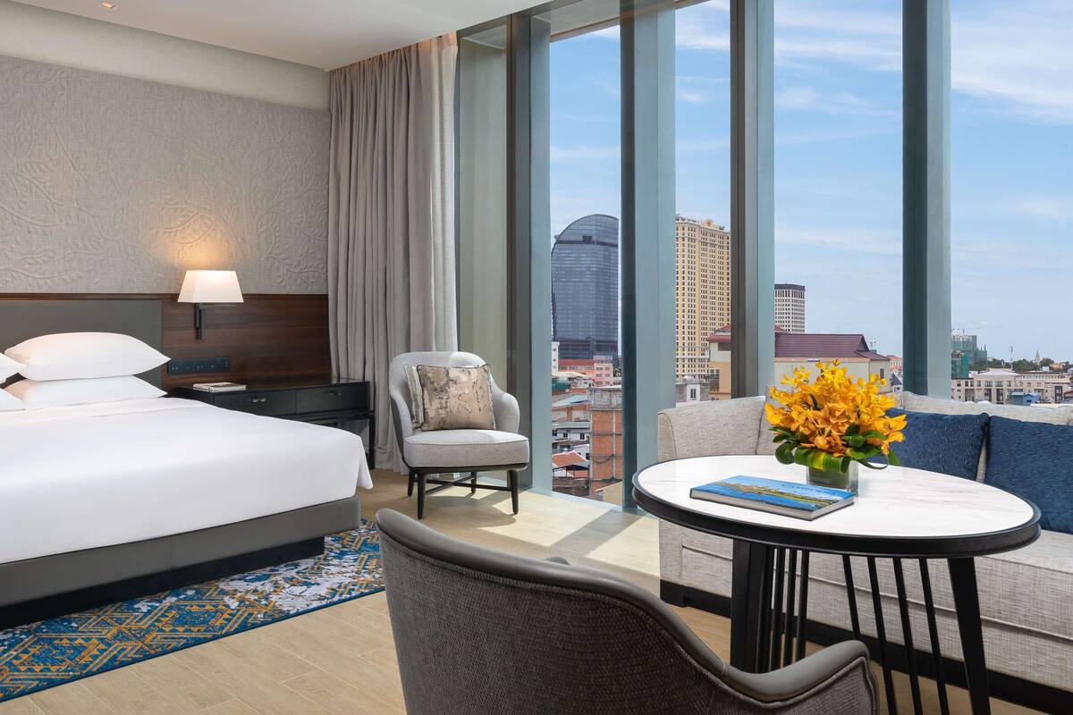 A hotel room with a bed, a chair, a table, and a view of the cityscape.