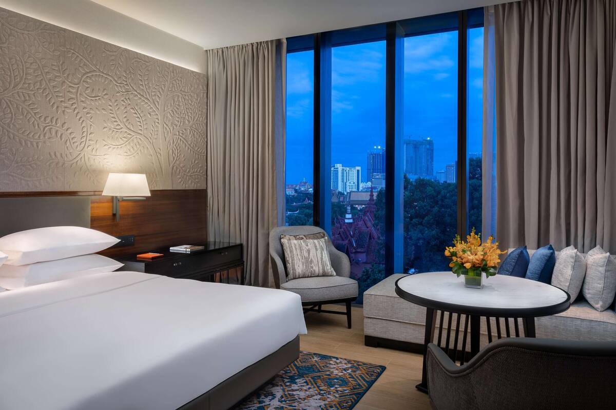 A hotel room with a large bed, a desk with a lamp, a chair, a sofa, and a view of a cityscape through large windows.