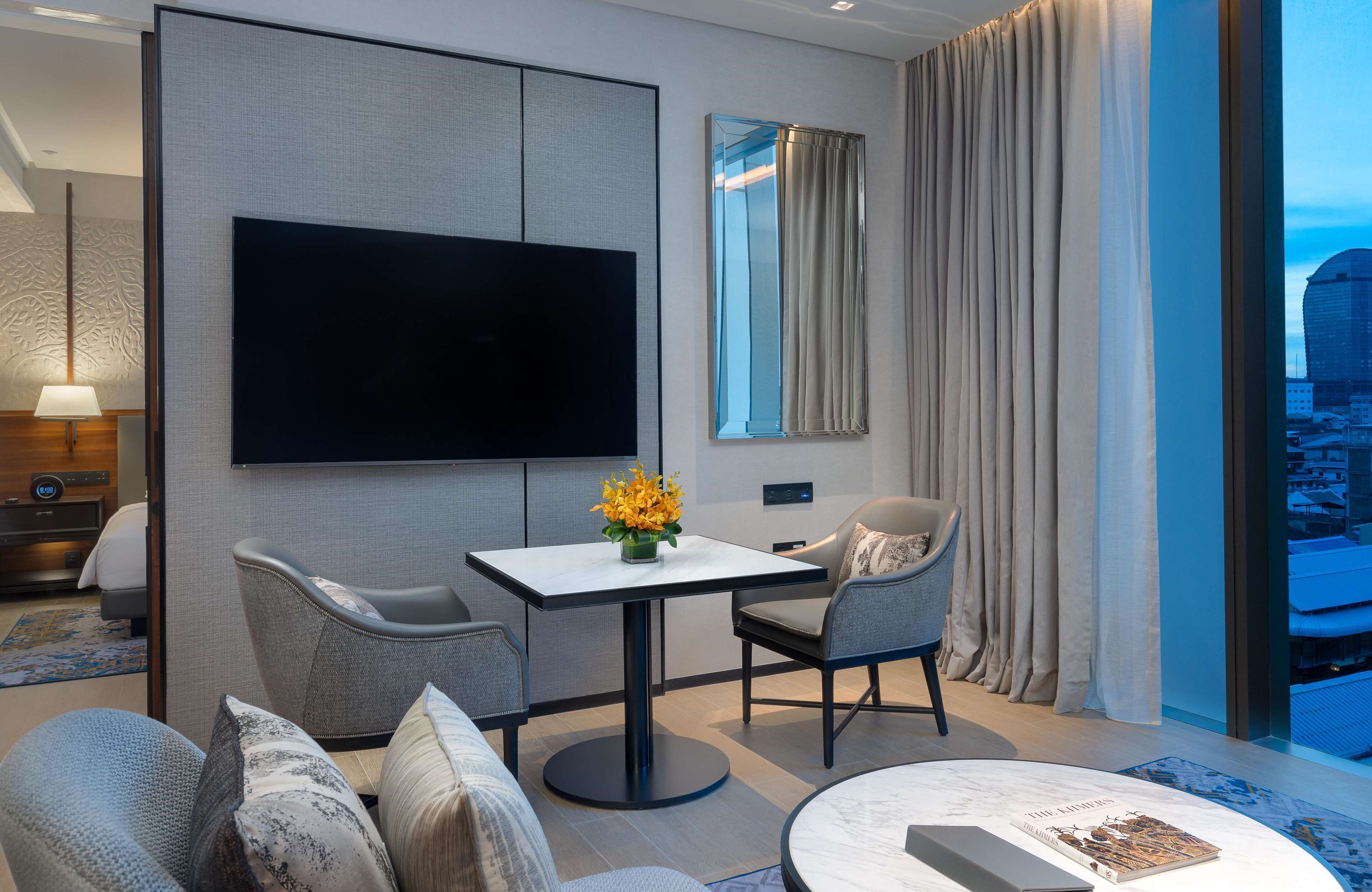 A modern hotel room with a flat-screen TV, a dining table with chairs, a sofa, a coffee table, and a large window with curtains.