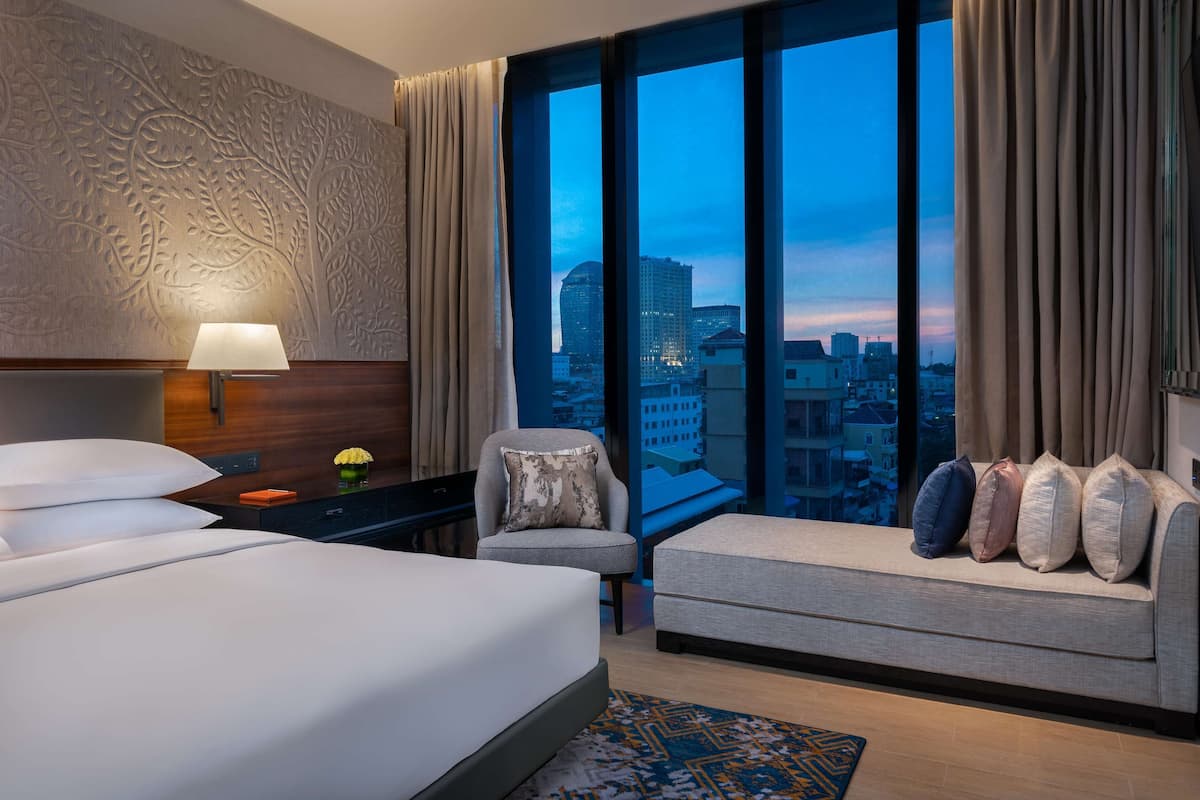 A hotel room with a large bed, a sofa with multiple pillows, a chair, a nightstand with a lamp, and a view of the cityscape through large windows.