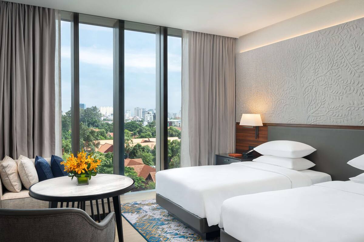 A hotel room with a large window, two beds, a desk, a chair, and a lamp.