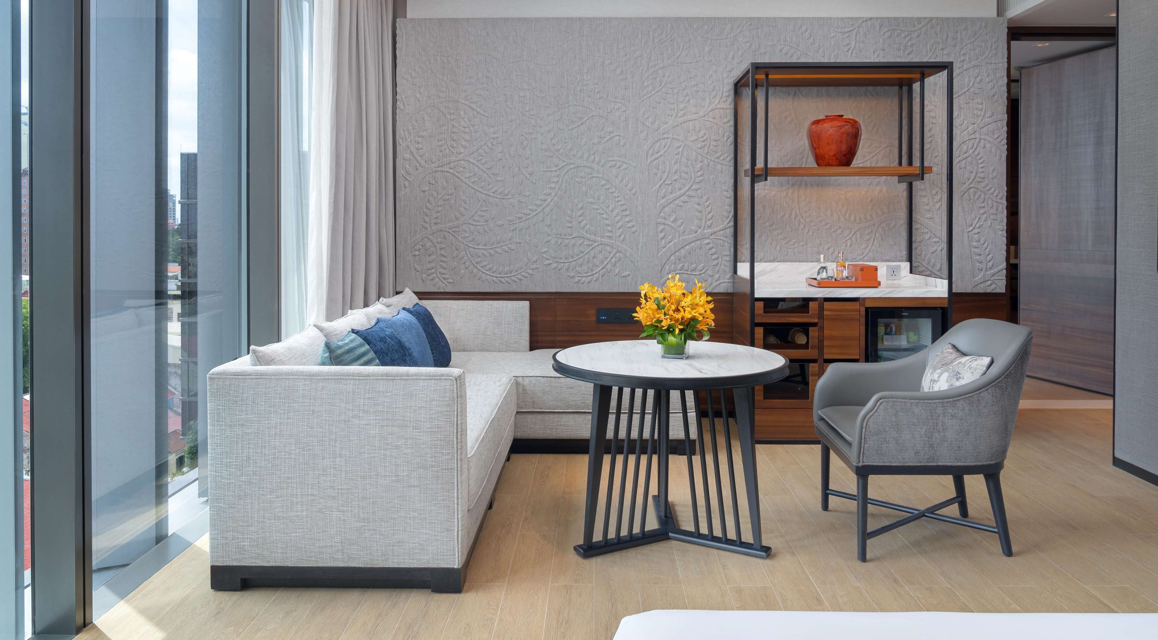 A modern hotel room with a sectional sofa, a small round table with a vase of flowers, a grey armchair, and a built-in shelf with a vase.