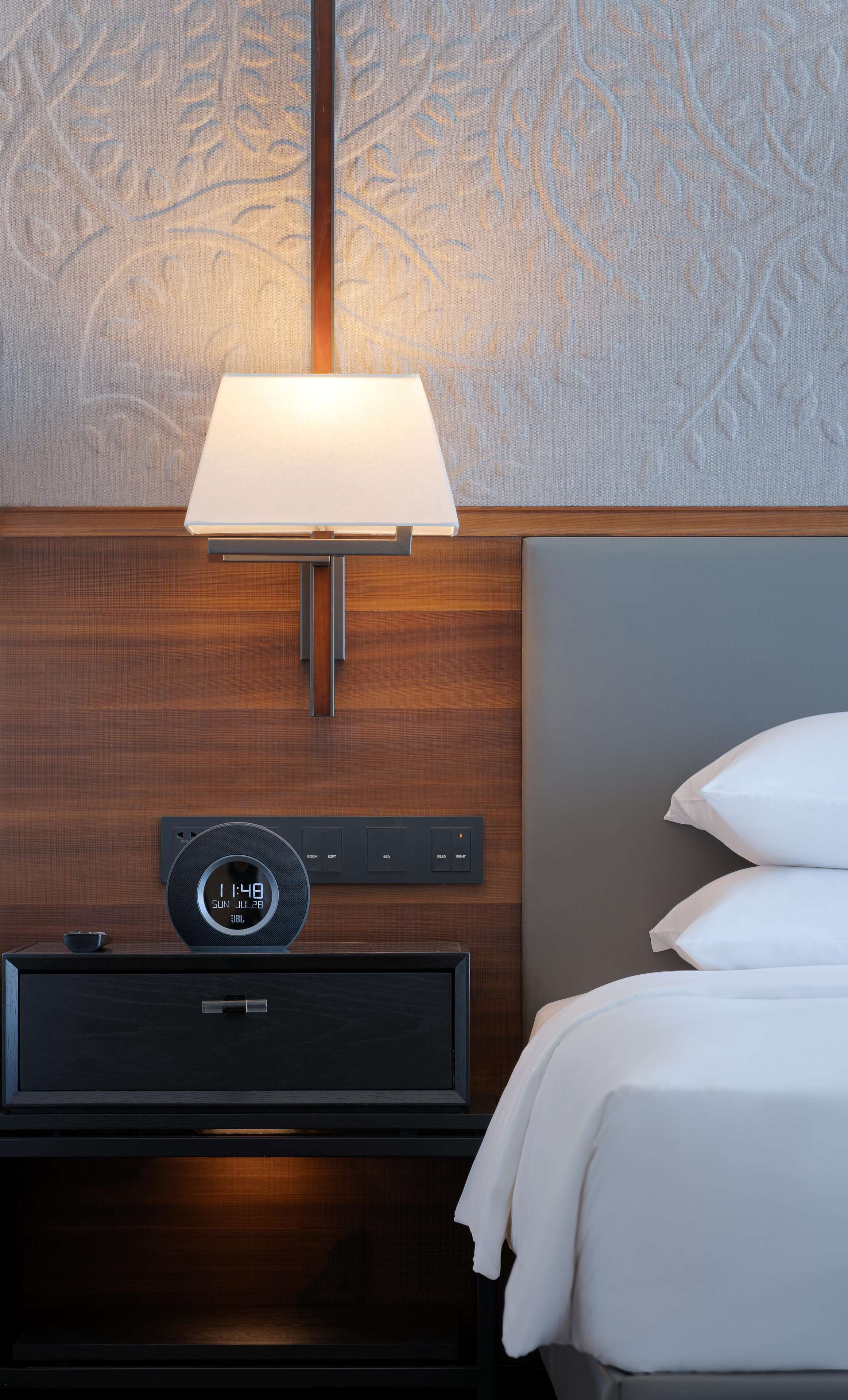 A hotel room with a bed, a bedside table with a clock, a lamp, and a wall with a leaf pattern.