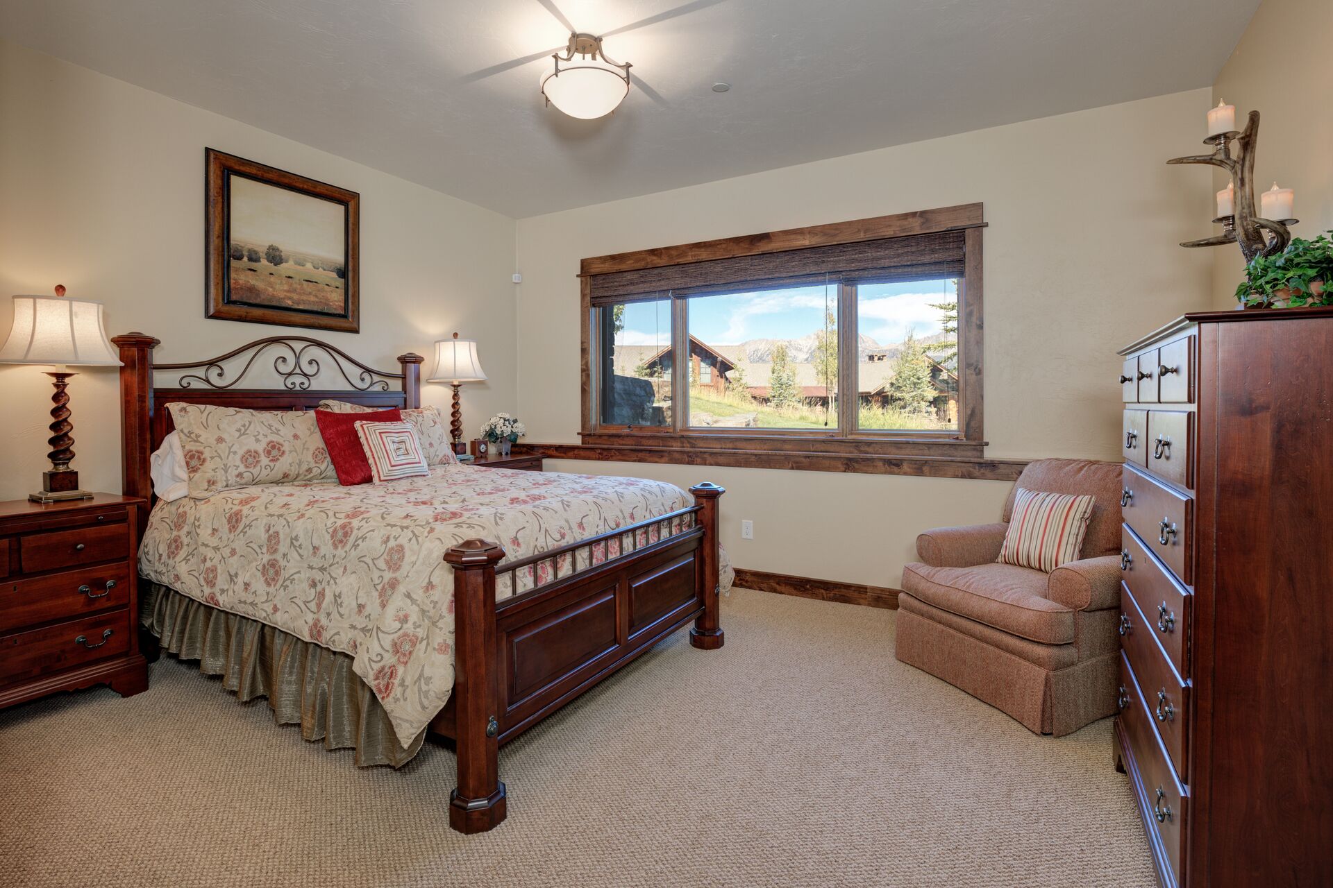 A bedroom with a wooden bed, two bedside tables with lamps, a chair, a dresser, and a window with a view of houses and mountains.